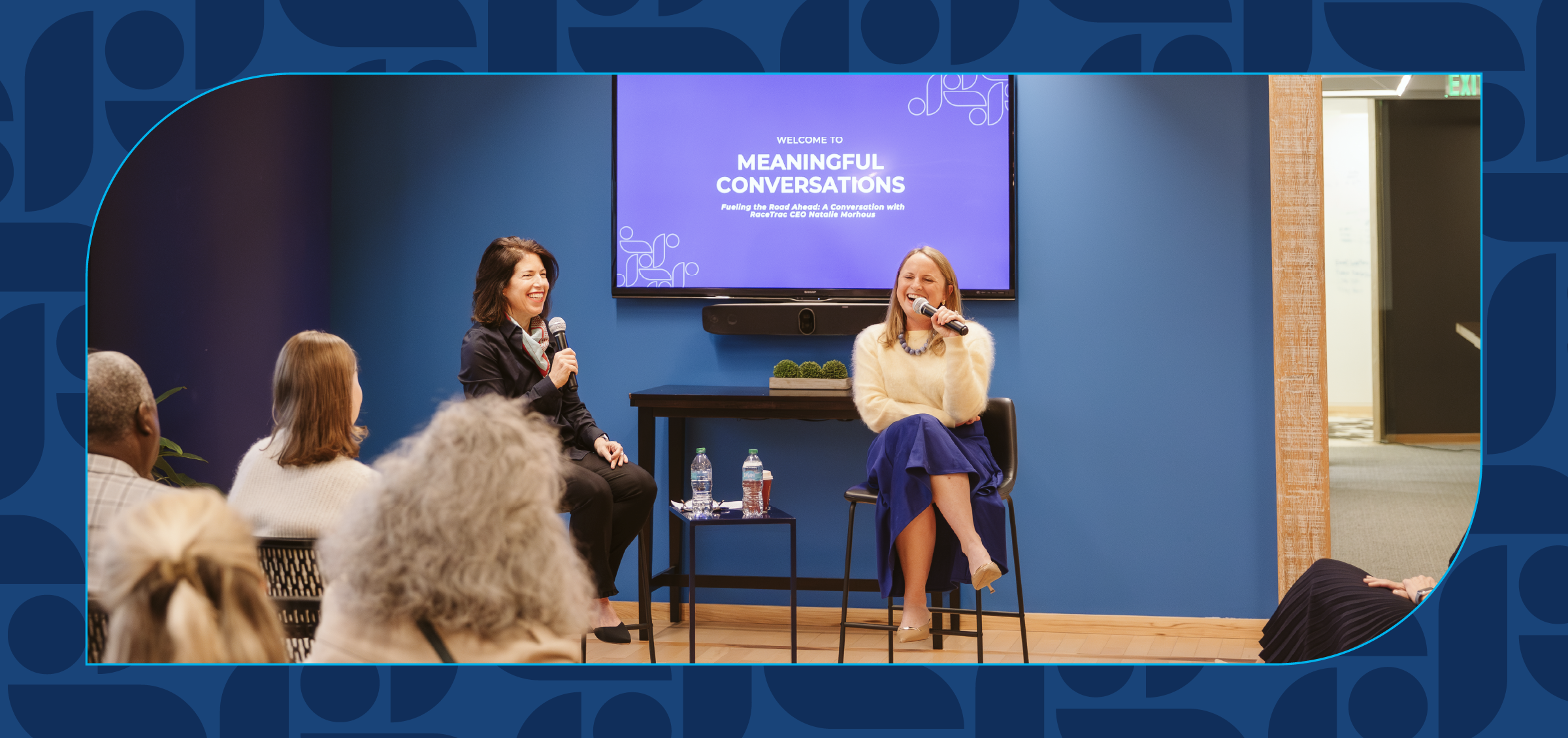 Two women sit on stage in a panel setup, smiling and speaking into microphones, with an audience in front and a purple screen behind reading 'Meaningful Conversations'.
