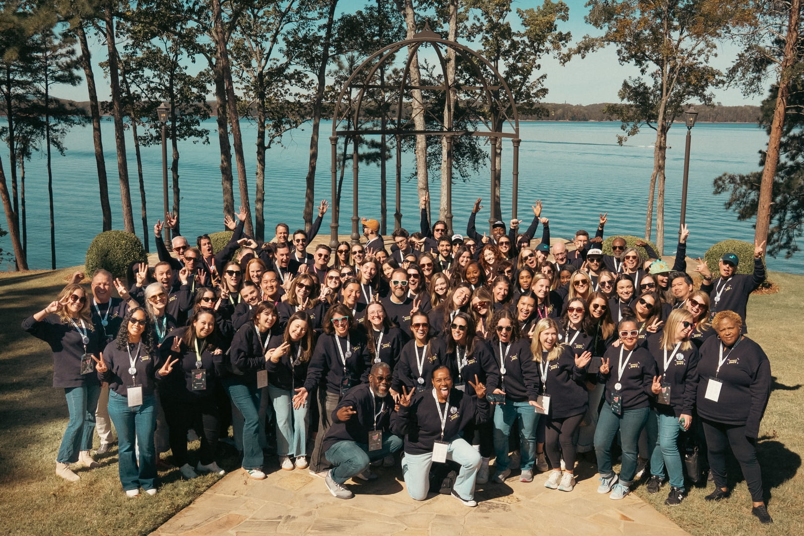 Large group of people posing together outdoors by a lake, wearing matching dark shirts and lanyards, many flashing peace signs.